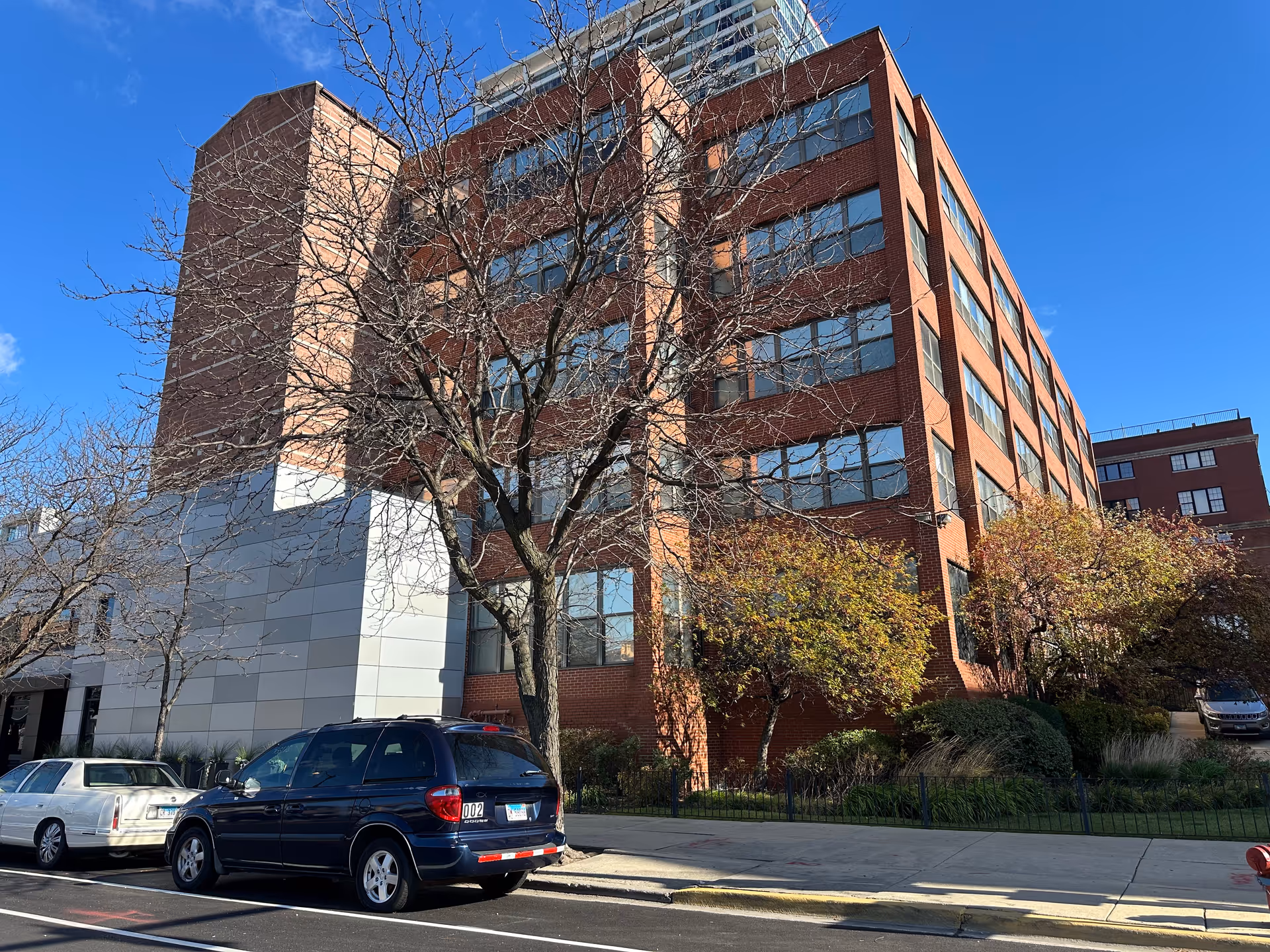 Red brick multi-story senior living building with parked cars and trees along the sidewalk under a blue sky.