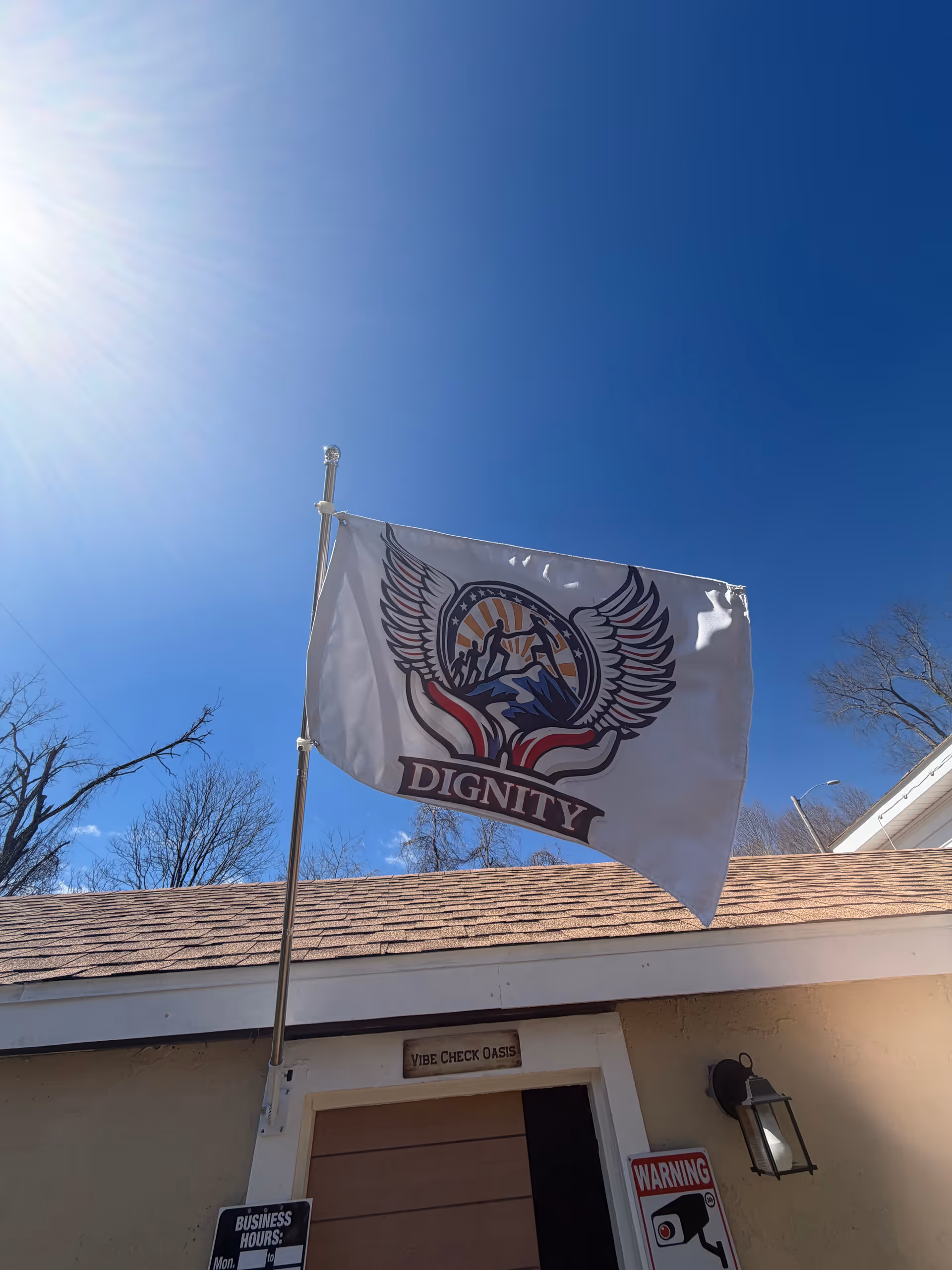 Flag with Dignity emblem on a pole in front of a small building and blue sky.