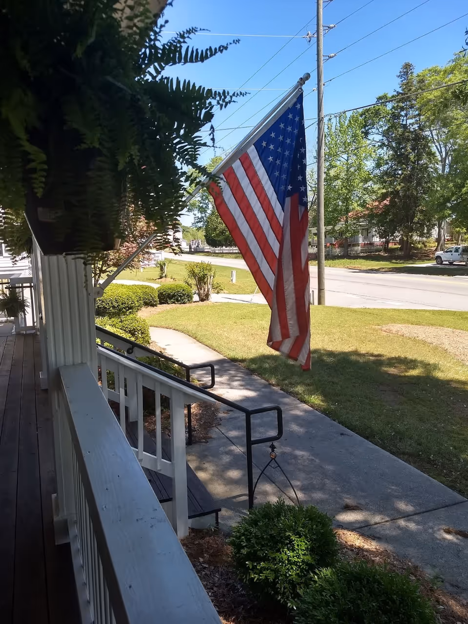View from a front porch showing an American flag, white railing, a walkway, and lawn leading to the street.