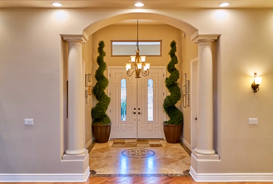 Elegant interior foyer with double entry doors, columns, chandelier, and tall potted topiary plants.