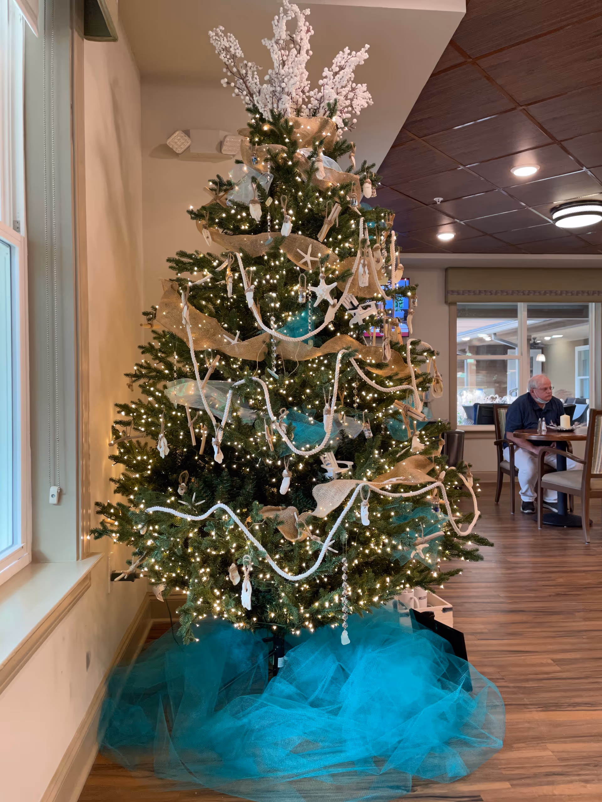Decorated Christmas tree in a bright common area with a resident seated nearby.