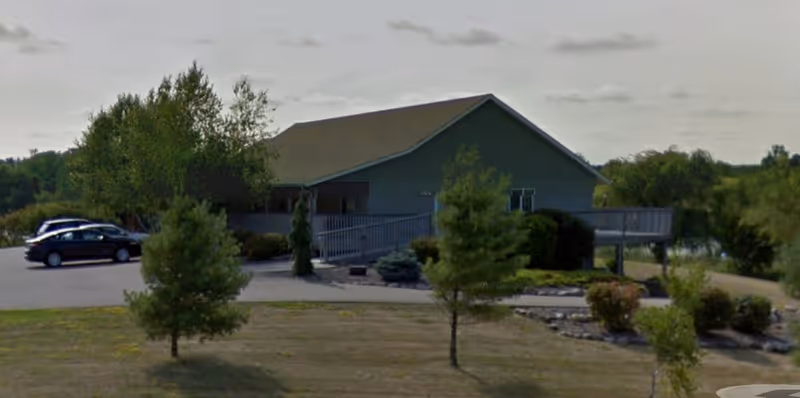 Single-story senior living facility exterior with ramped entrance, parking lot, and surrounding trees.