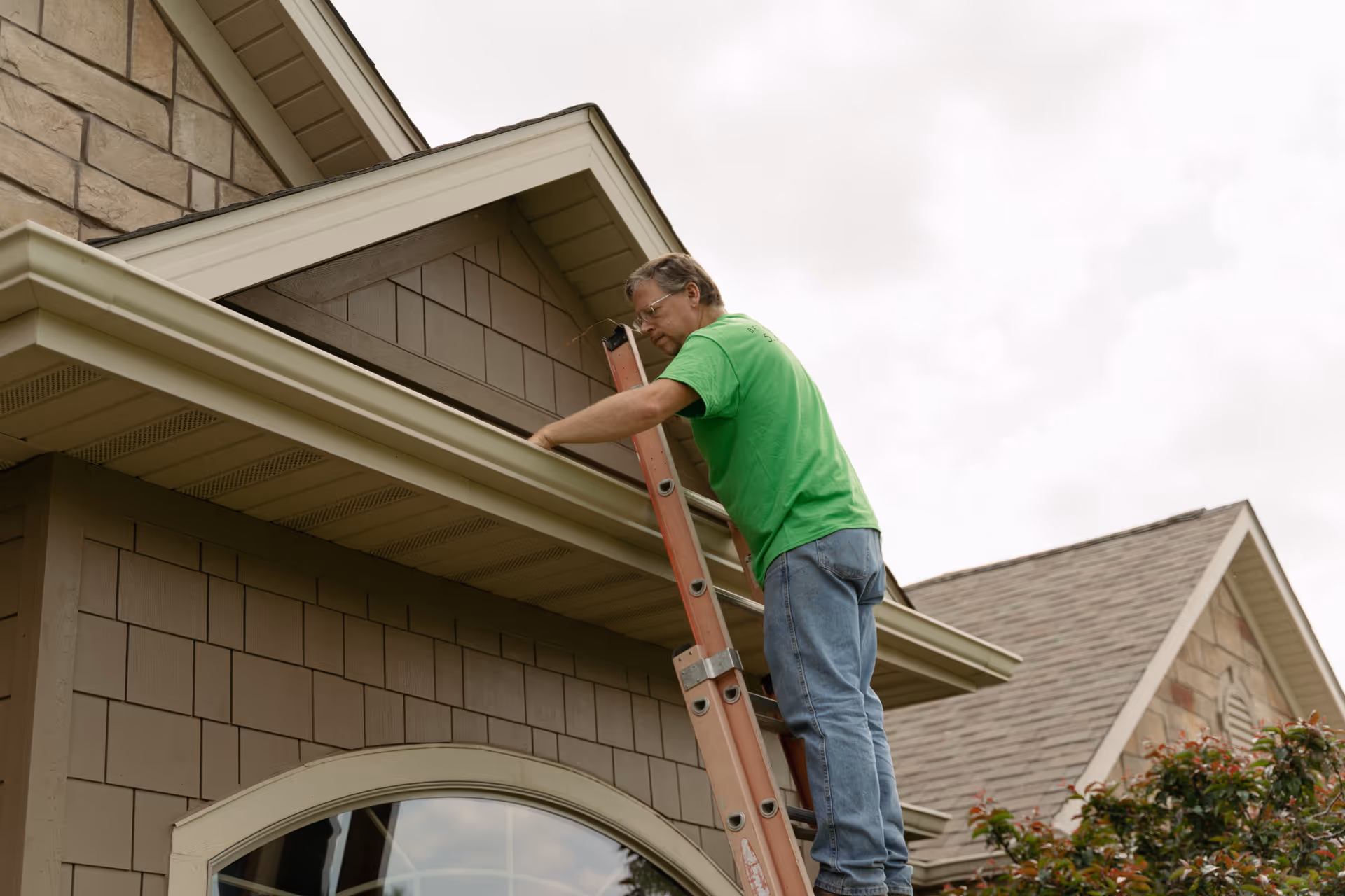 A man on a ladder repairs the eaves of a suburban house exterior.