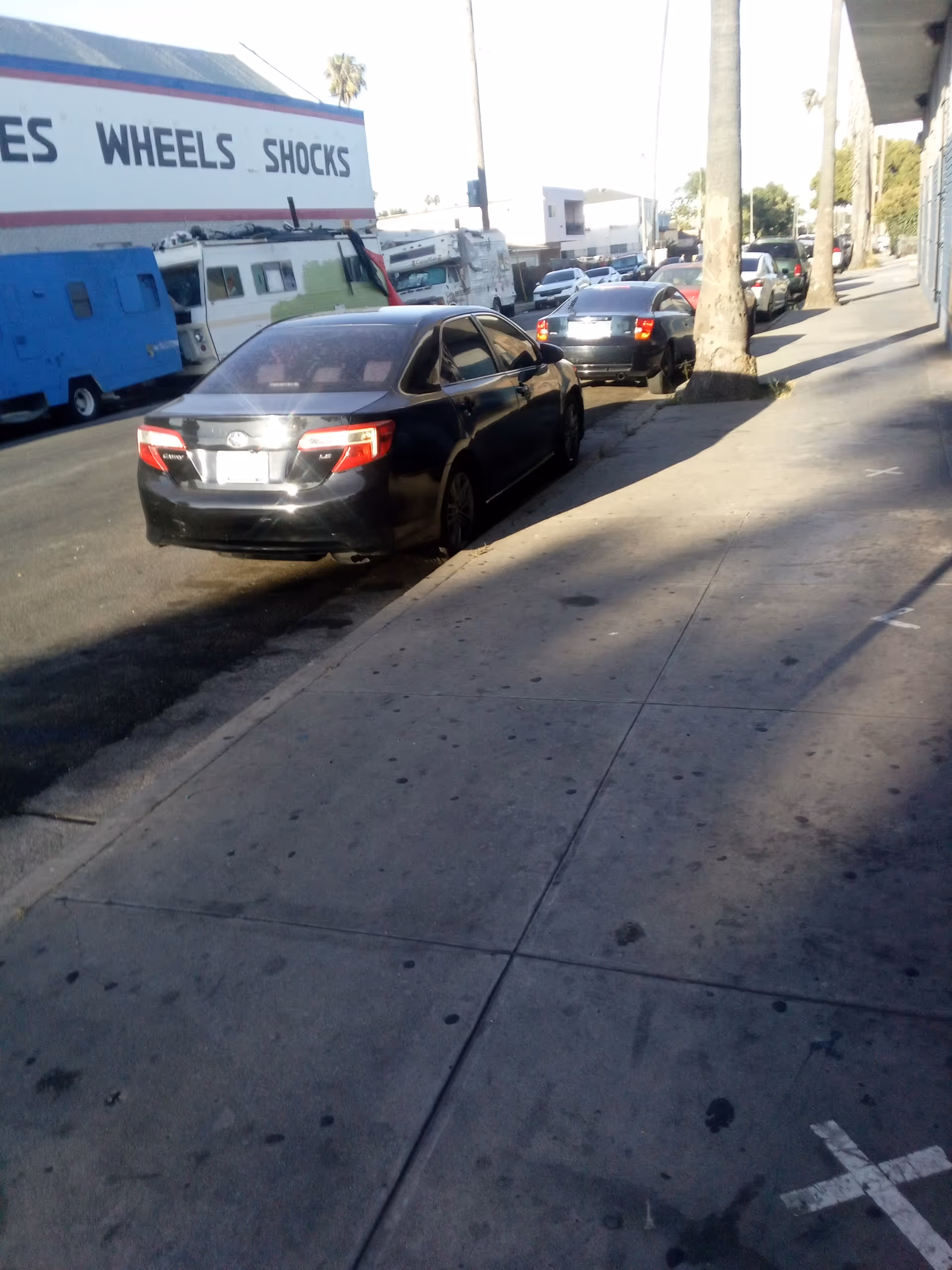 Outdoor city street with parked cars beside a storefront trailer displaying a large WHEELS SHOCKS sign.