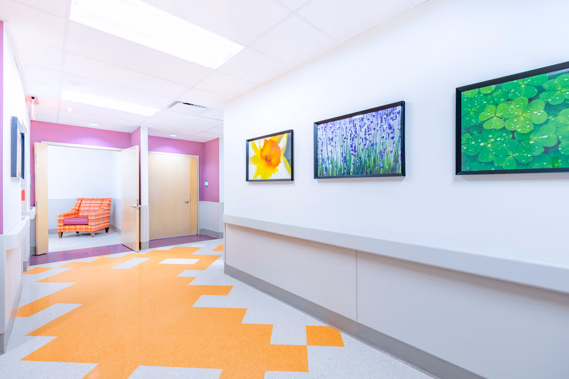 A brightly lit hallway with white walls and a patterned orange and gray floor. On the right wall, there are three framed pictures featuring a yellow flower, purple flowers, and green leaves. At the end of the hallway, there are two doors, one open revealing an orange and red plaid armchair inside a room with purple walls.