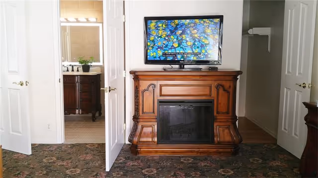 A room with floral carpet and a wooden fireplace mantel with a flat-screen TV on top. Two white doors are open on either side of the fireplace, one leading to a bathroom with a vanity and mirror, and the other leading to a hallway.