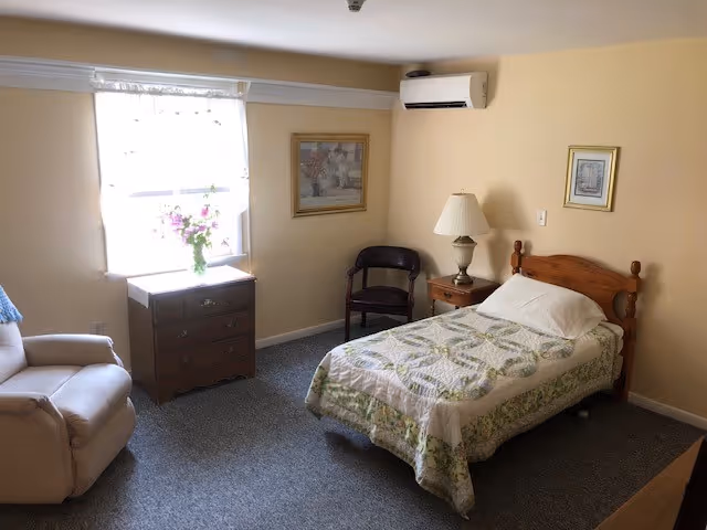 Single bedroom with bed, dresser, chair, lamp, window, and vase of flowers.