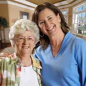 Two smiling women, one resident and a caregiver, stand together in a sunny living area.