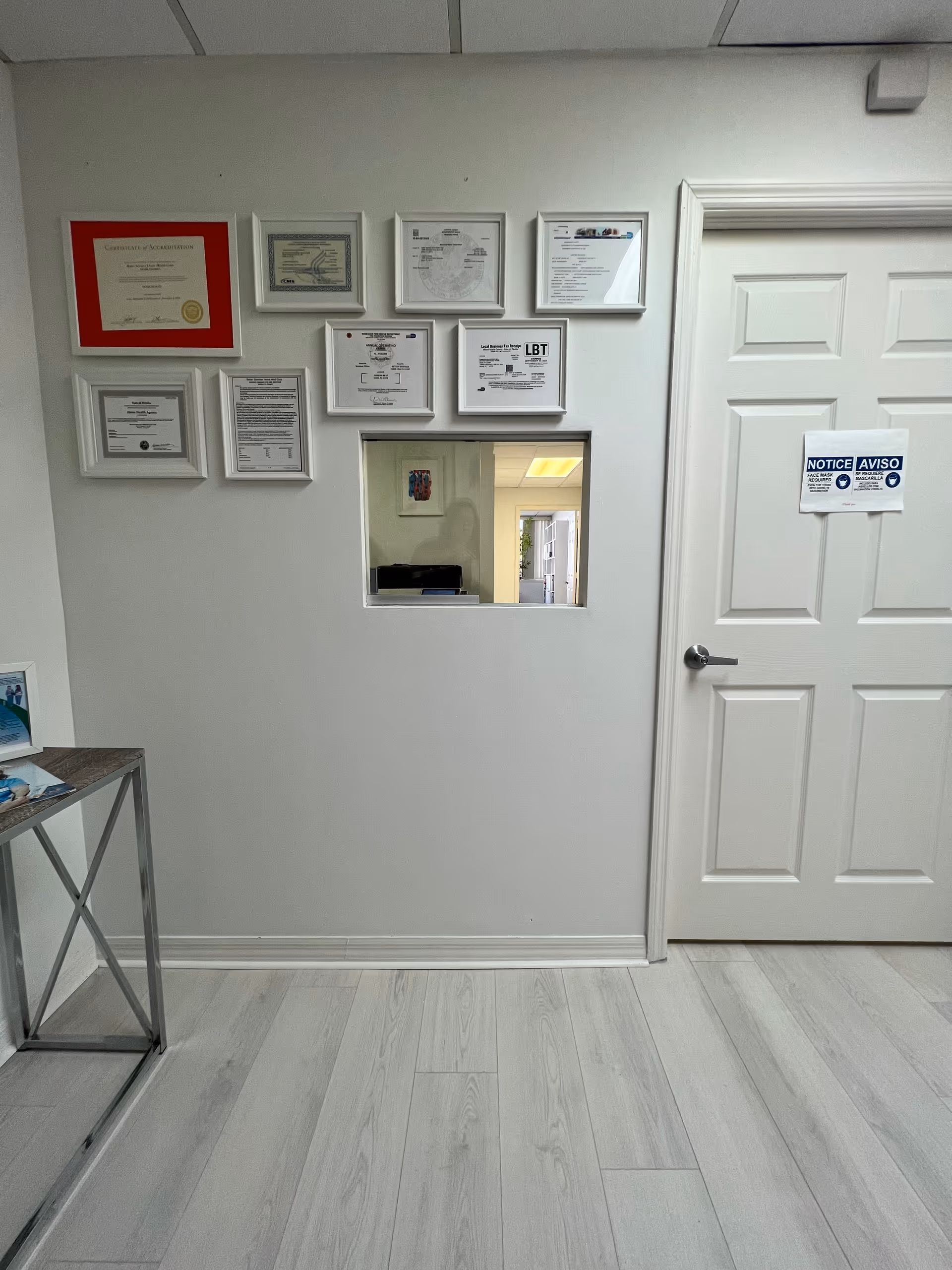 White wall with framed certificates, a small window into another room, door, and a metal table.