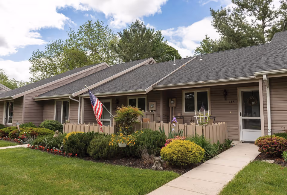 Front exterior of a single-story senior living townhouse with a small fenced porch, American flag, and landscaped lawn.