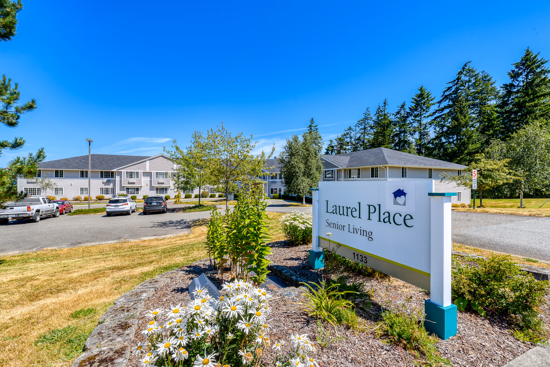 Exterior view of Laurel Place Senior Living facility on a sunny day with a clear blue sky. The image shows a large building with multiple windows, a parking lot with several cars, landscaped greenery, and a prominent sign in the foreground that reads 'Laurel Place Senior Living 1133'.