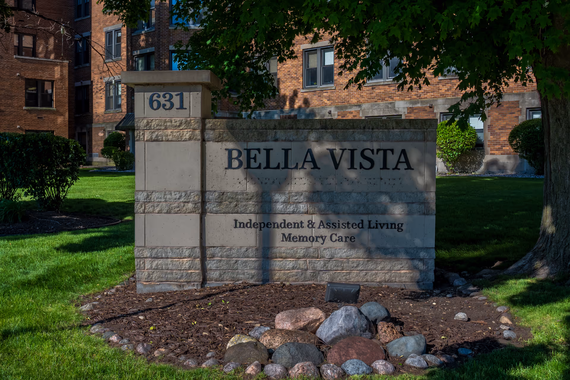 Stone entrance sign reading "BELLA VISTA" with address "631" and "Independent & Assisted Living Memory Care" in front of a brick building and landscaped lawn.