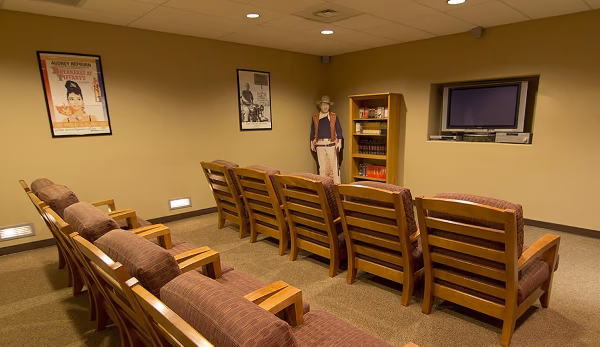 A senior living lounge with rows of wooden chairs facing a wall mounted TV and posters.