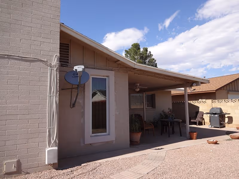 Outdoor patio area of a single-story building with a covered porch, a satellite dish mounted on the wall, a table with chairs, potted plants, a barbecue grill, and a gravel ground with a stone pathway under a partly cloudy sky.