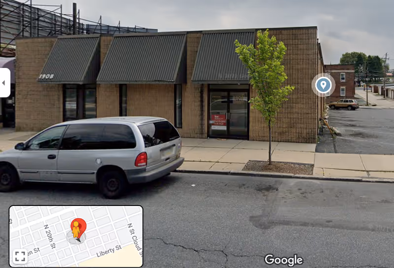 Brick storefront with black awnings, sidewalk, and silver van parked outside.