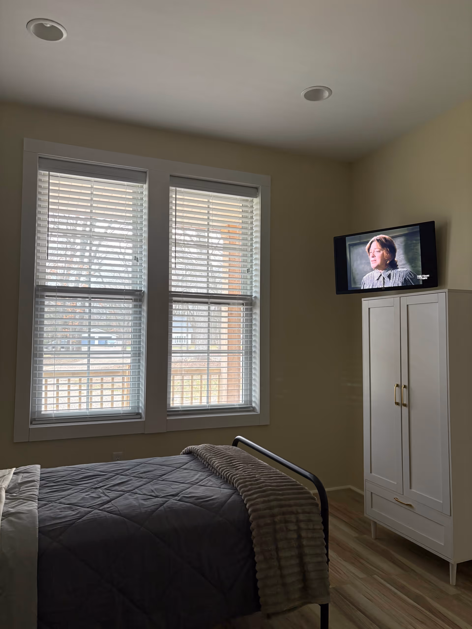 A bedroom with a bed, blinds, and a wall-mounted TV on a white cabinet.