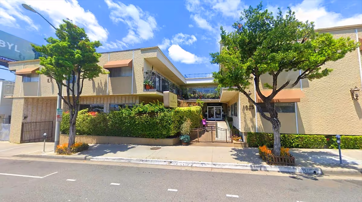 Two-story assisted living building with beige exterior, trees, hedges, and a central entrance.
