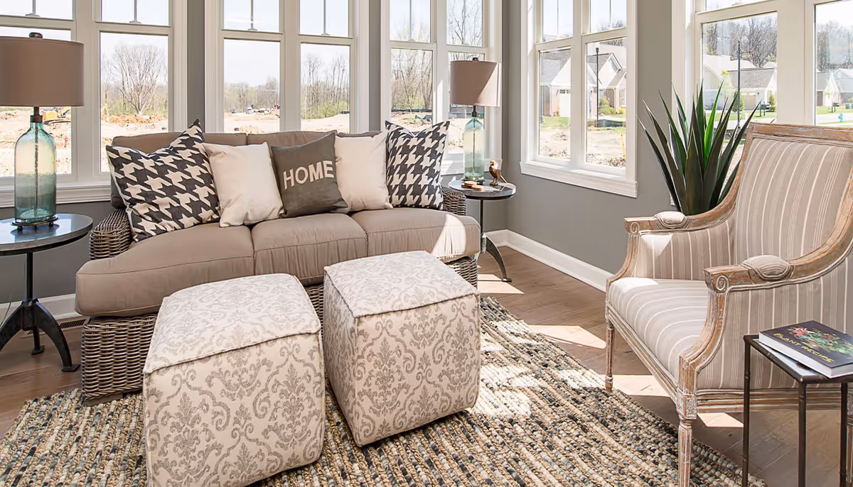 A bright living room with large windows letting in natural light. The room features a beige sofa with decorative pillows, including one with the word 'HOME', two patterned ottomans, a striped armchair, a woven rug, two side tables with glass lamps, and a tall green plant near the window.