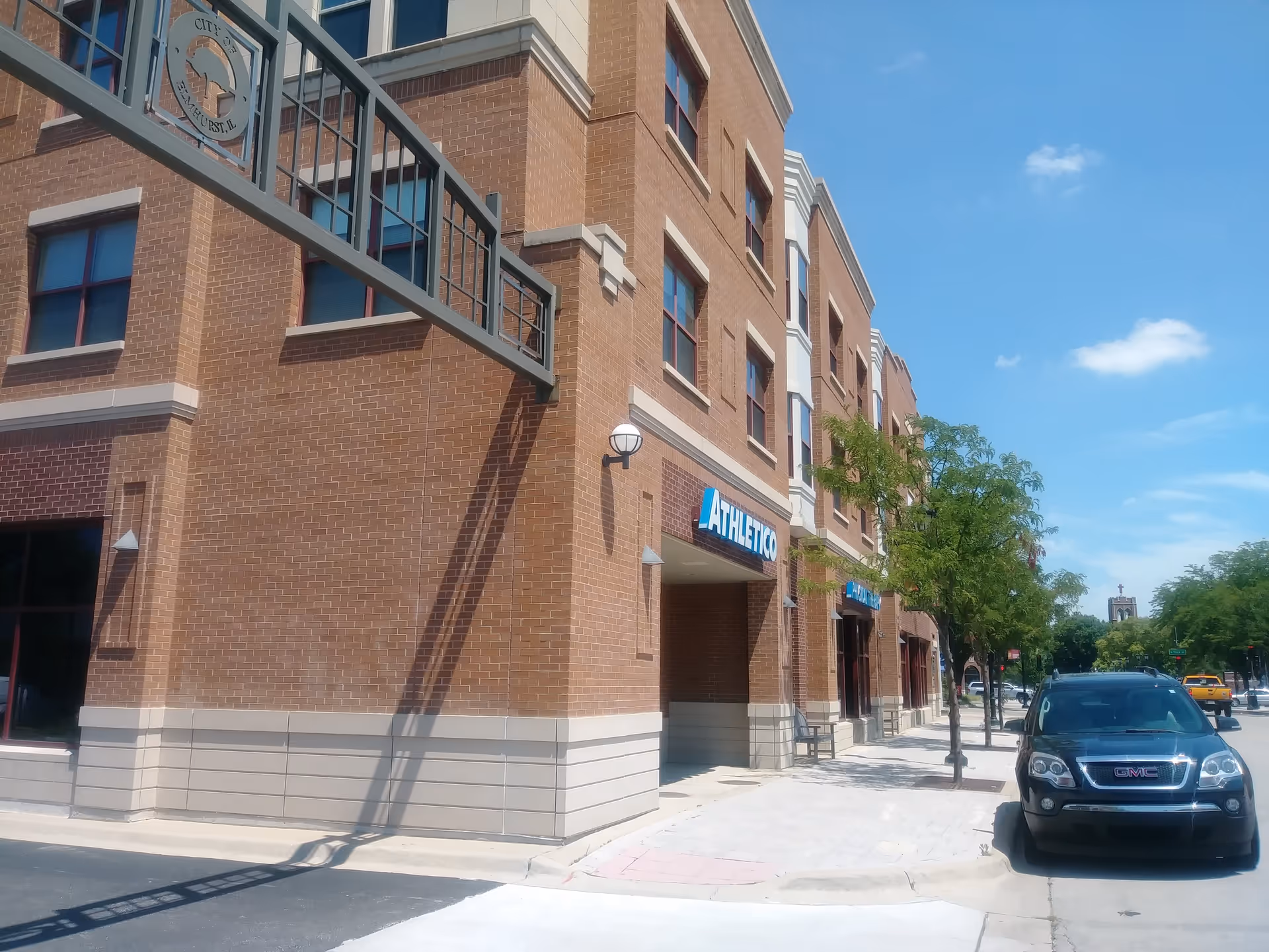 Brick multi-story building along a sidewalk with trees and a parked SUV.