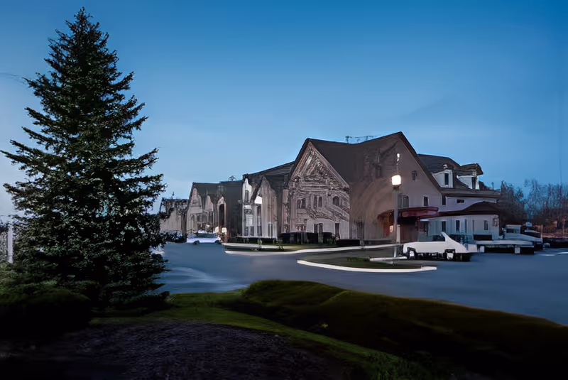 Exterior view of a large senior living facility building at dusk with a tall evergreen tree in the foreground and a curved driveway leading to the entrance.