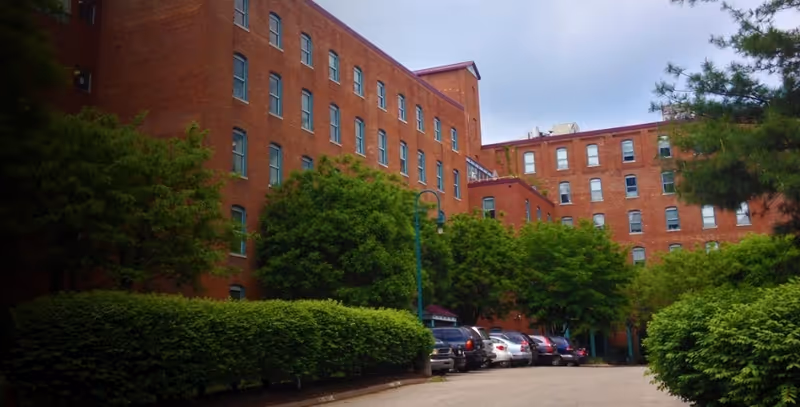 Red brick senior living facility exterior with trees and parked cars along the curb.