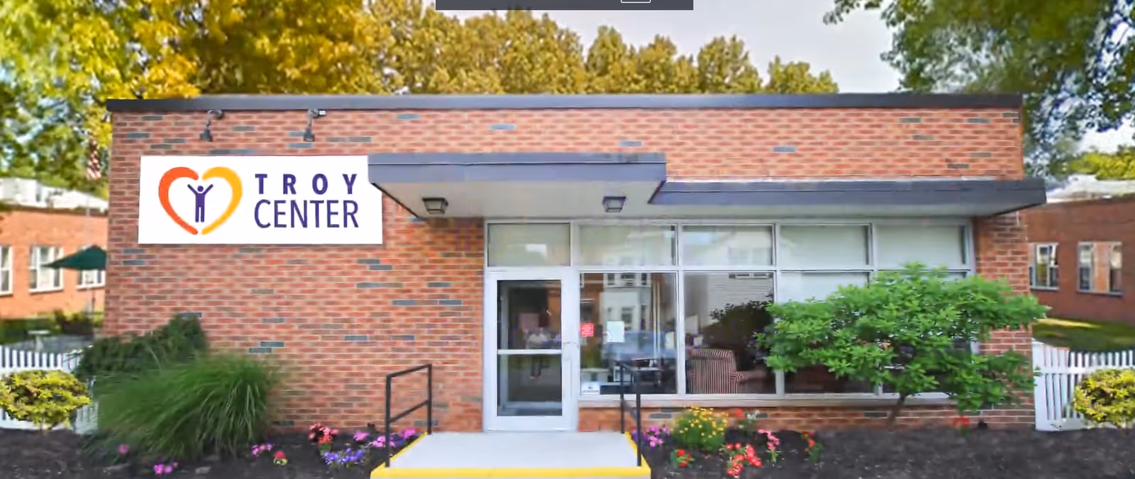 Front exterior view of a single-story brick building with a sign that reads 'Troy Center'. The entrance has a glass door and large windows, with a small ramp and stairs leading up to it. There are bushes, flowers, and trees surrounding the building.