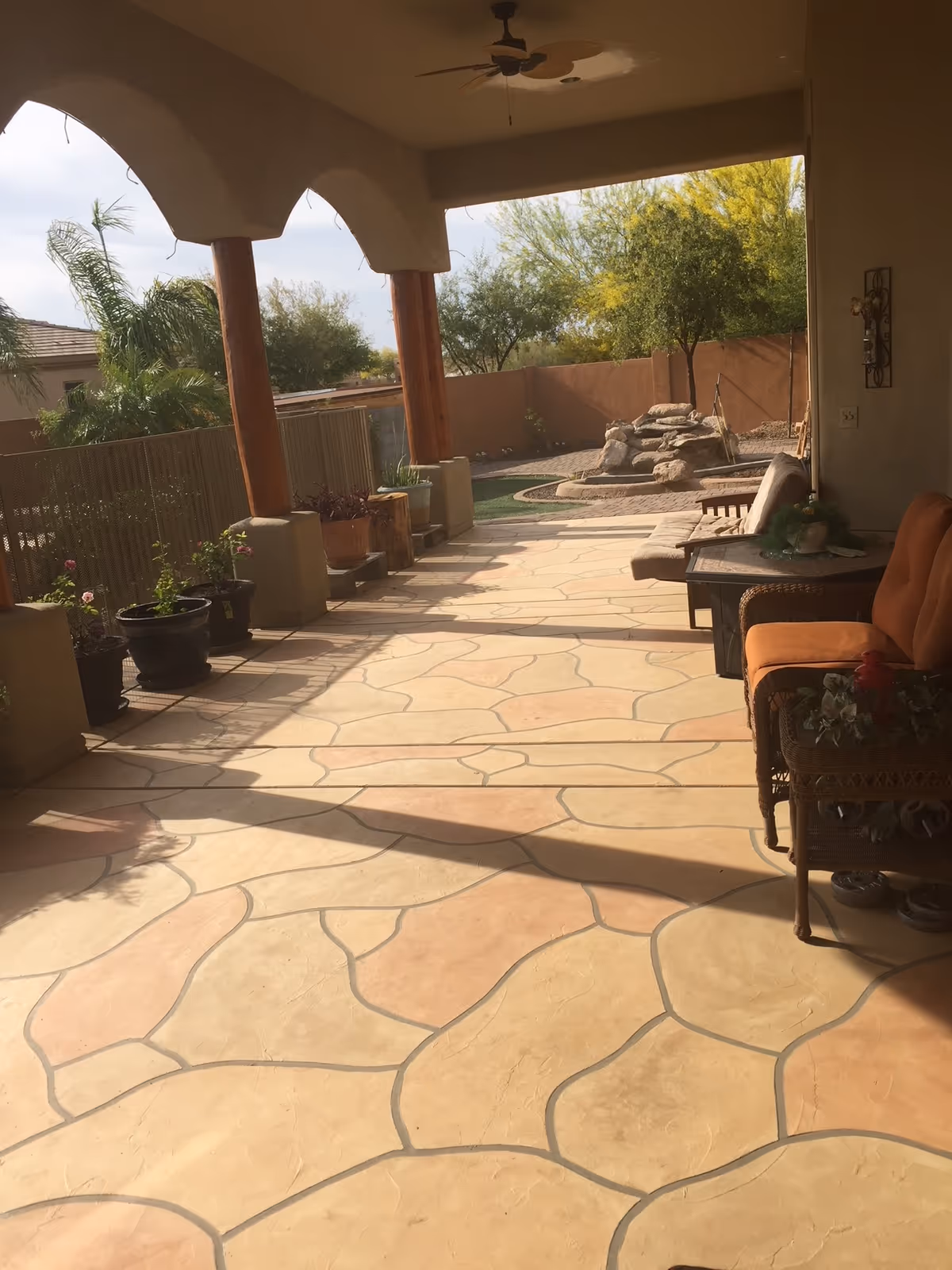 Covered patio with patterned stone-style flooring, wooden support columns, potted plants, outdoor seating, and a rock water feature in the backyard.