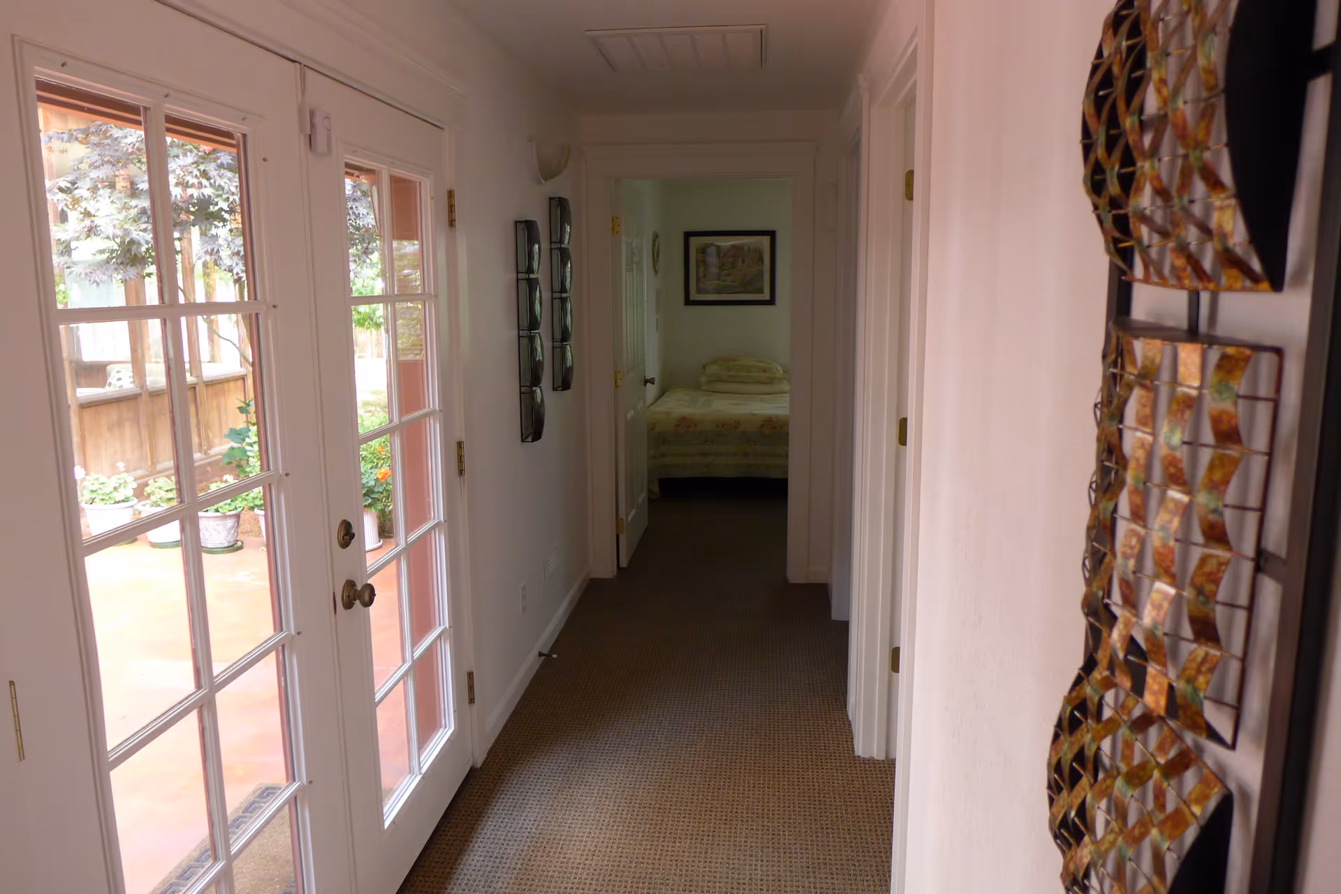 A hallway with carpeted floor leading to a bedroom at the end. The bedroom has a bed with floral bedding and a framed picture on the wall. On the left side of the hallway, there are glass double doors opening to an outdoor patio area with potted plants and trees. The right wall of the hallway features decorative metal wall art.