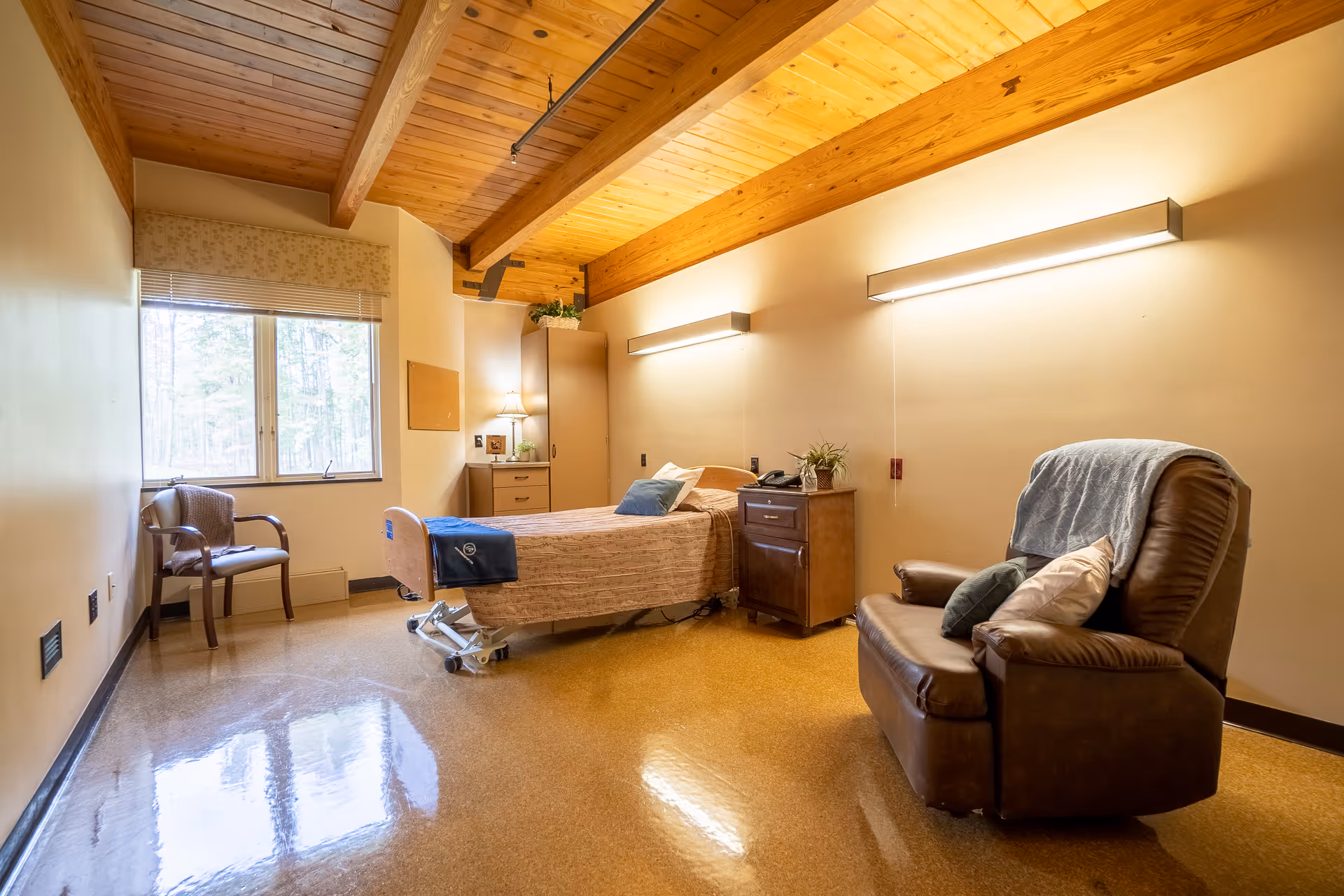 A well-lit assisted living or memory care bedroom with a single hospital-style bed covered with a beige blanket and a blue throw. Next to the bed is a wooden nightstand with a plant and a telephone. A comfortable brown recliner chair with pillows and a blanket is positioned nearby. The room features a wooden ceiling with exposed beams, a window with a view of trees, a wooden cabinet, and a chair near the window.