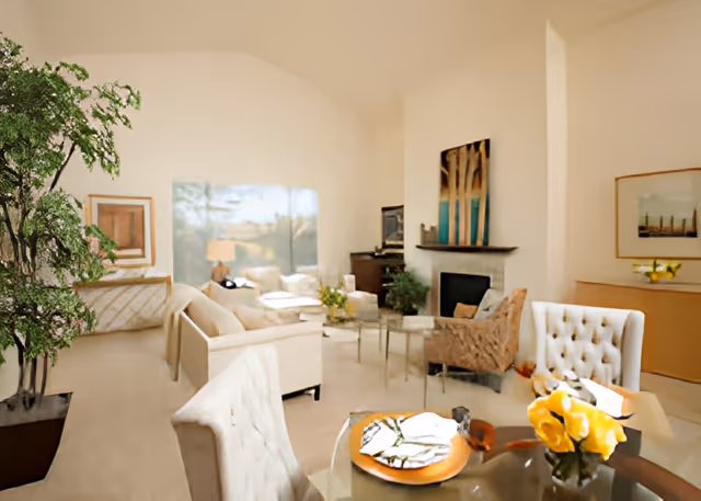 A bright and spacious living room with beige walls and carpet. The room features multiple comfortable chairs and sofas arranged around a glass coffee table with a flower vase. There is a fireplace with artwork above it, a large window letting in natural light, and a potted plant in the corner. A dining table with tufted chairs and a centerpiece of yellow flowers is visible in the foreground.