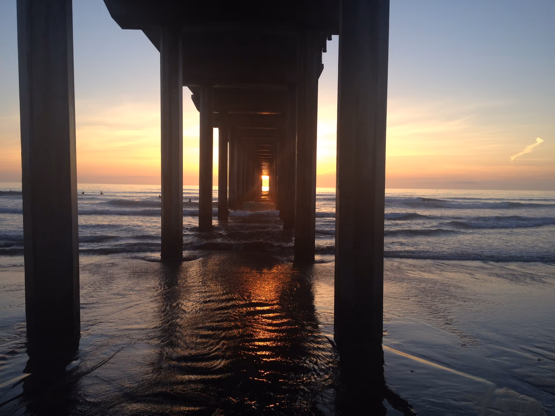 Sunset spills through a pier's columns onto the wet beach and waves.