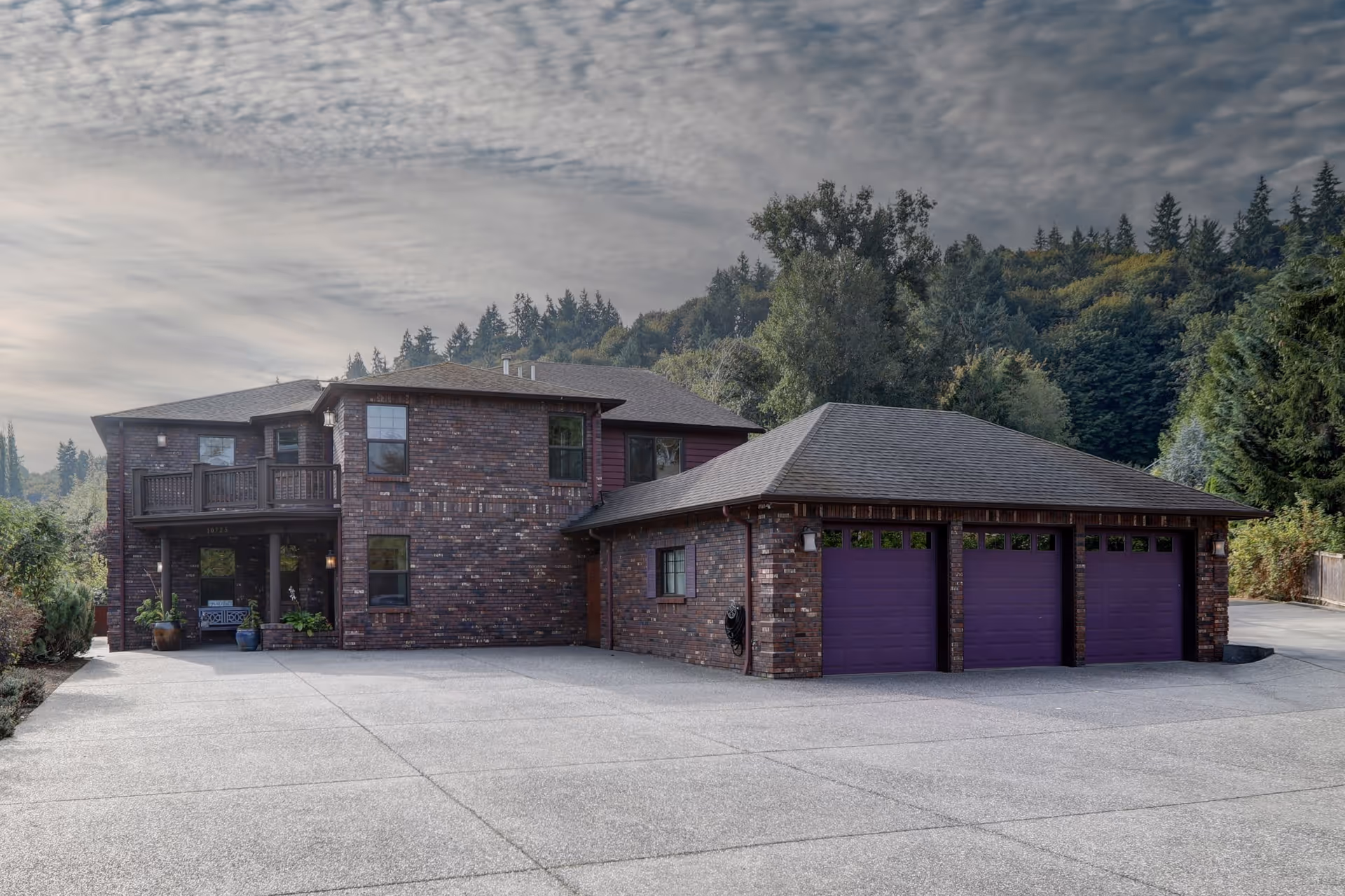 Exterior view of a two-story brick building with a three-car garage featuring purple doors, surrounded by trees and greenery under a cloudy sky.