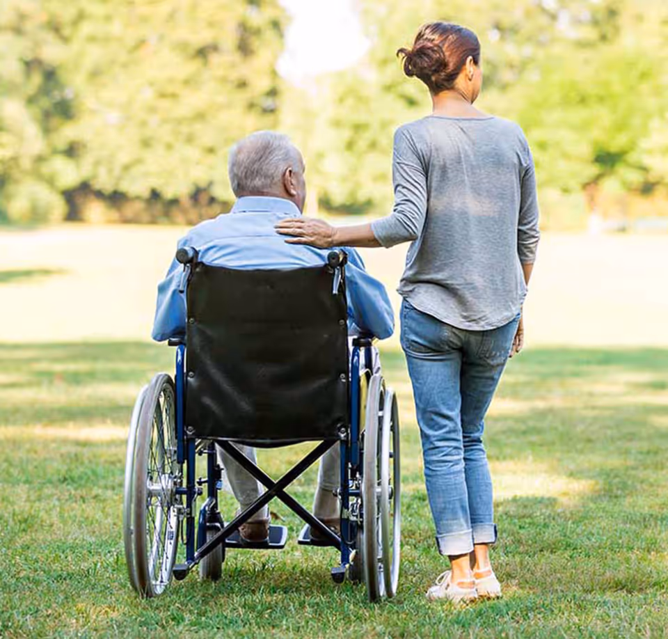 A caregiver walks beside an elderly man seated in a wheelchair in a park.