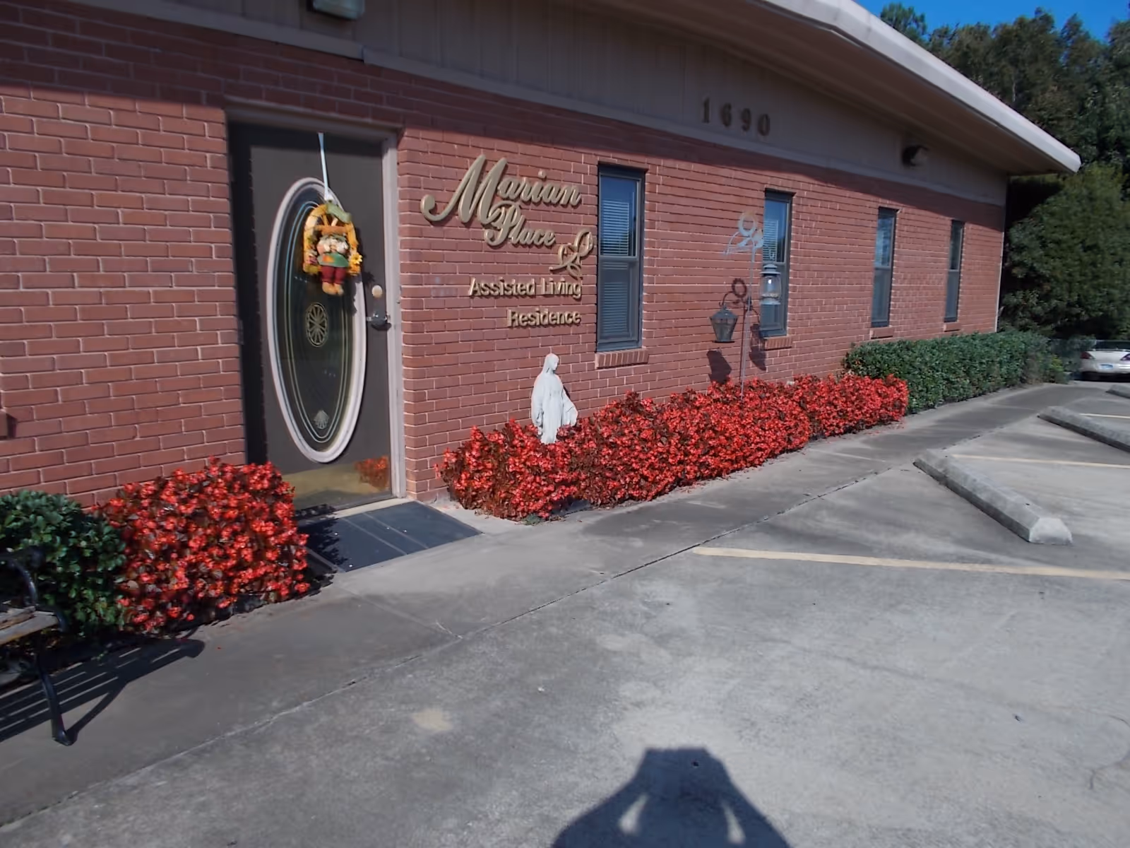 Exterior view of Marian Place Assisted Living Residence showing a brick building with a glass door decorated with a fall-themed wreath. There are red flowering bushes along the building and a small statue near the entrance. The building number 1690 is visible above the door, and there is a parking area in front.