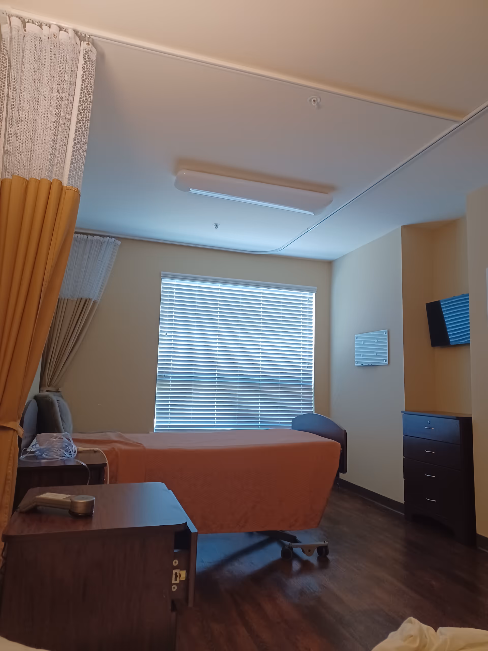 Single bedroom in senior living facility with orange bedspread, curtains, window blinds, and dresser.