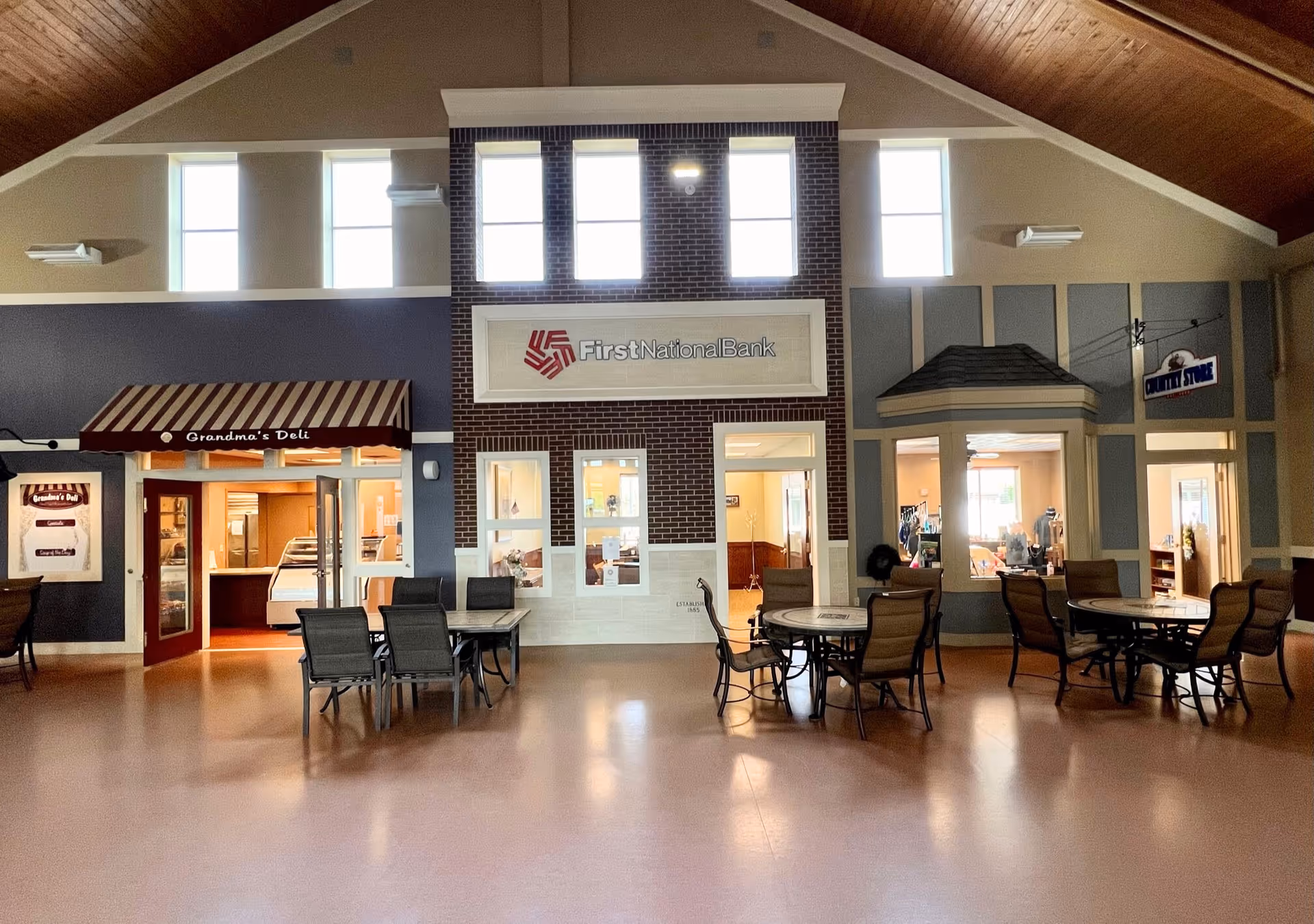 Interior common area with storefronts, tables, chairs, and a high vaulted ceiling.