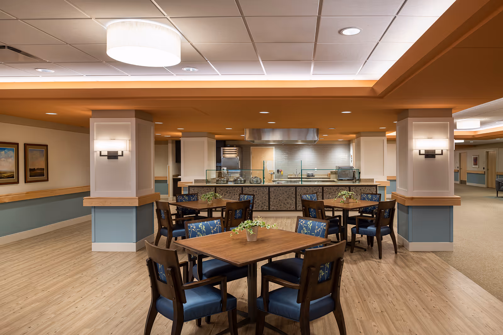 Bright dining room with wooden tables and blue upholstered chairs, open kitchen in background