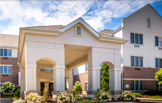 Covered portico entrance of a multi-story senior living building with columns and landscaped flowerbeds.