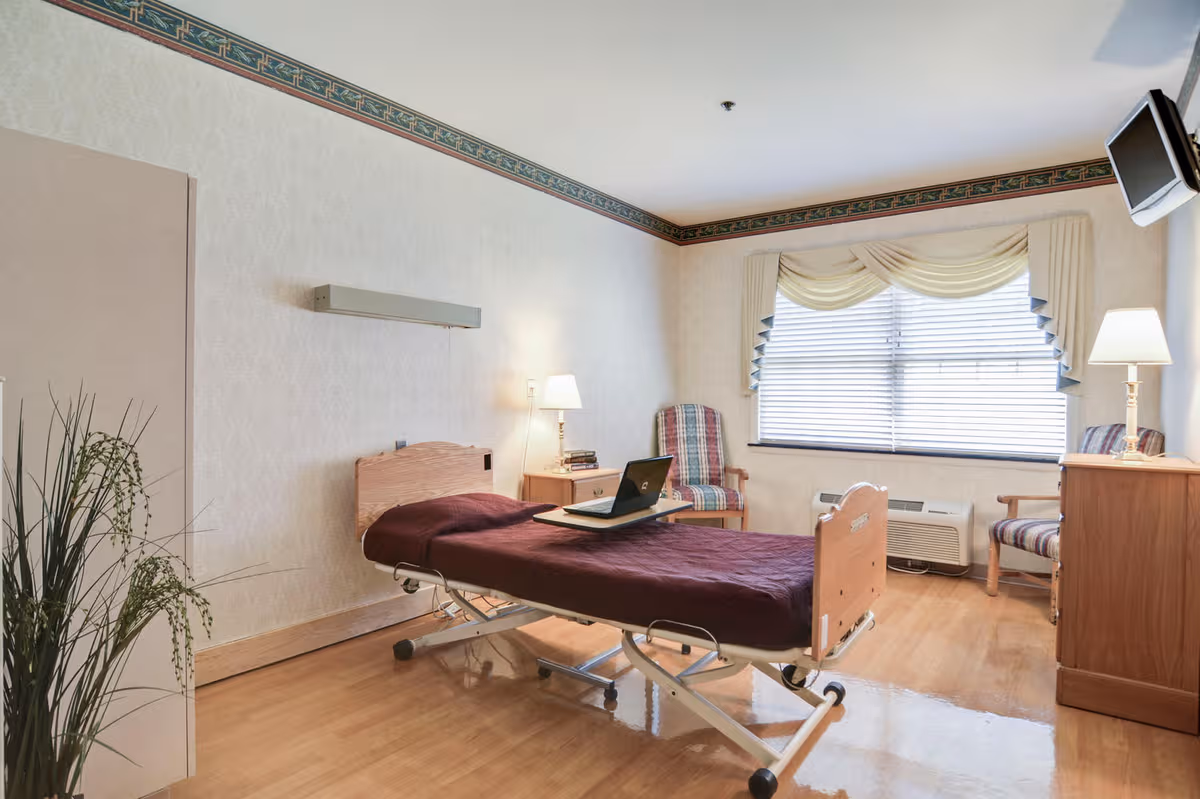 A senior care room with a hospital bed, desk, chairs, lamps, and a large window.