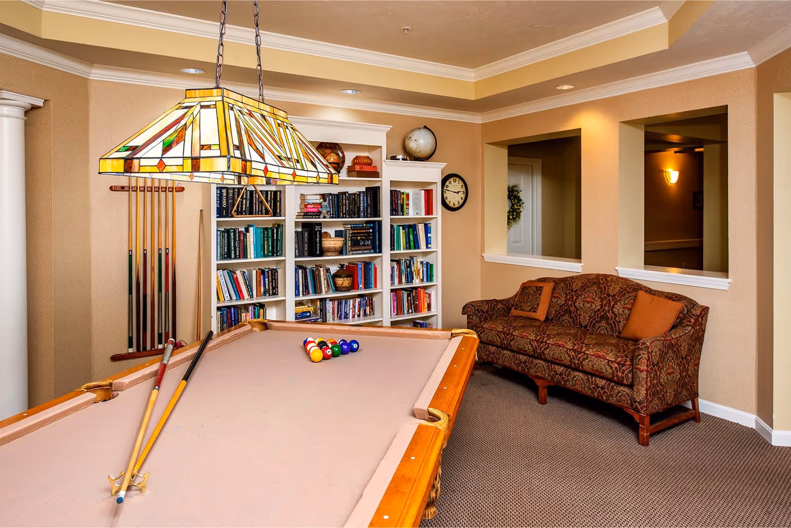 A cozy game room featuring a pool table with cues and balls arranged for a game. Behind the pool table is a white bookshelf filled with books and decorative items, including a globe. To the right, there is a patterned sofa with two orange cushions. The room has beige walls, carpeted floor, and a stained glass pendant light hanging above the pool table.
