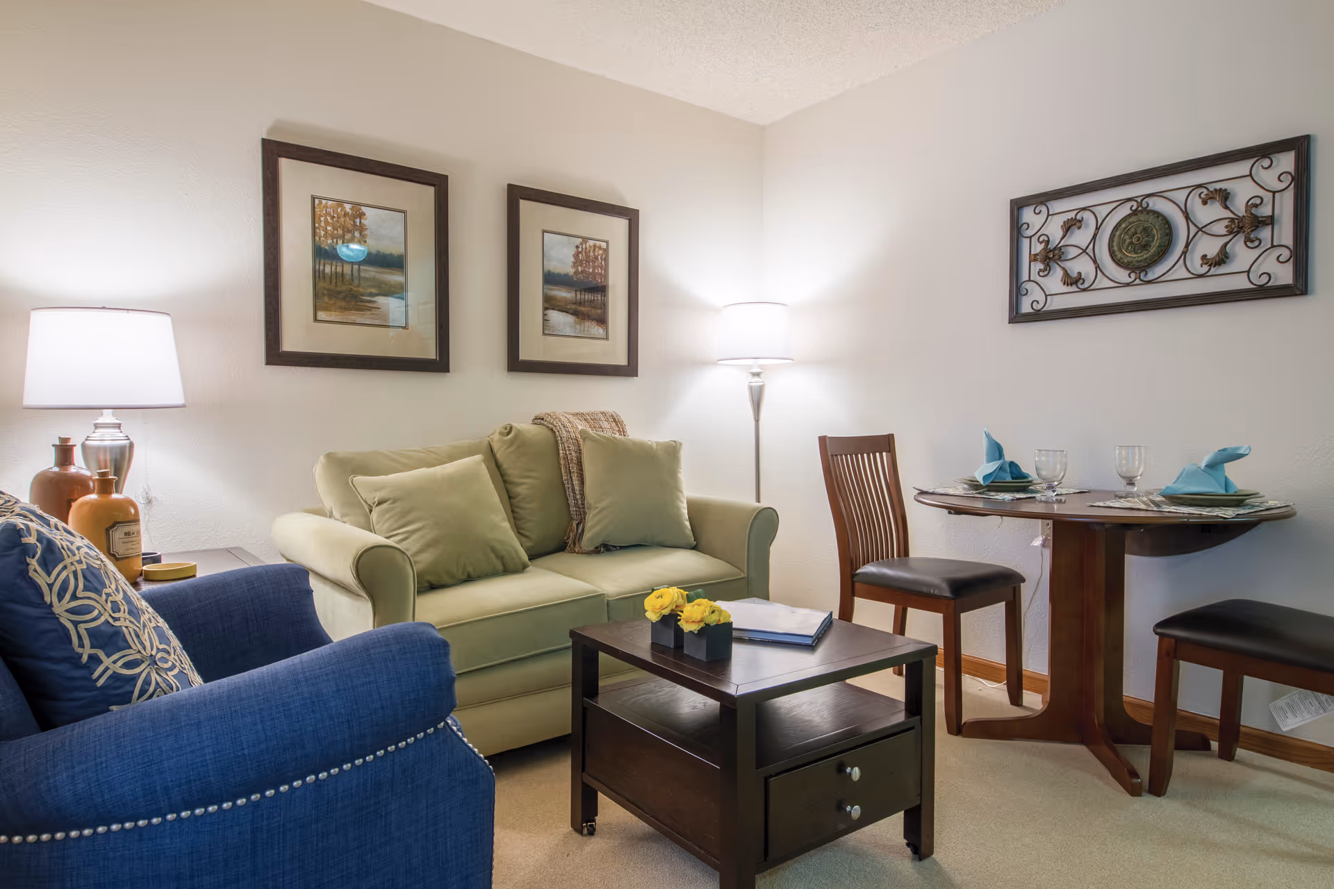 Cozy living room with a green loveseat, blue armchair, coffee table and a small round dining table set against a white wall with framed art and lamps.