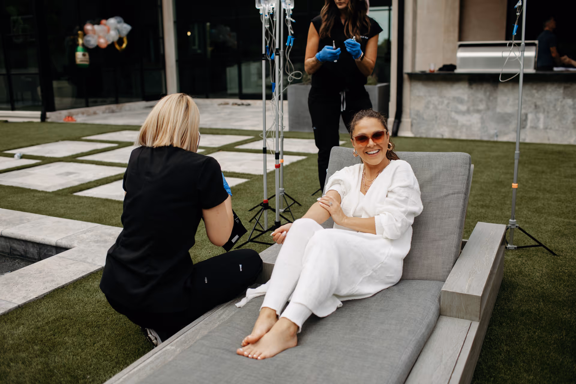Outdoor courtyard with a smiling woman in white receiving IV therapy from a nurse.