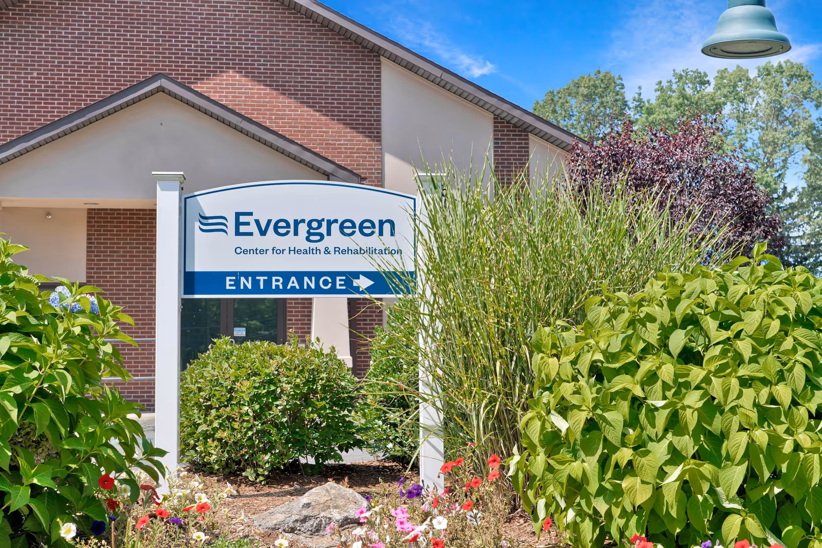 Evergreen entrance sign beside brick building framed by lush shrubs and flowers.