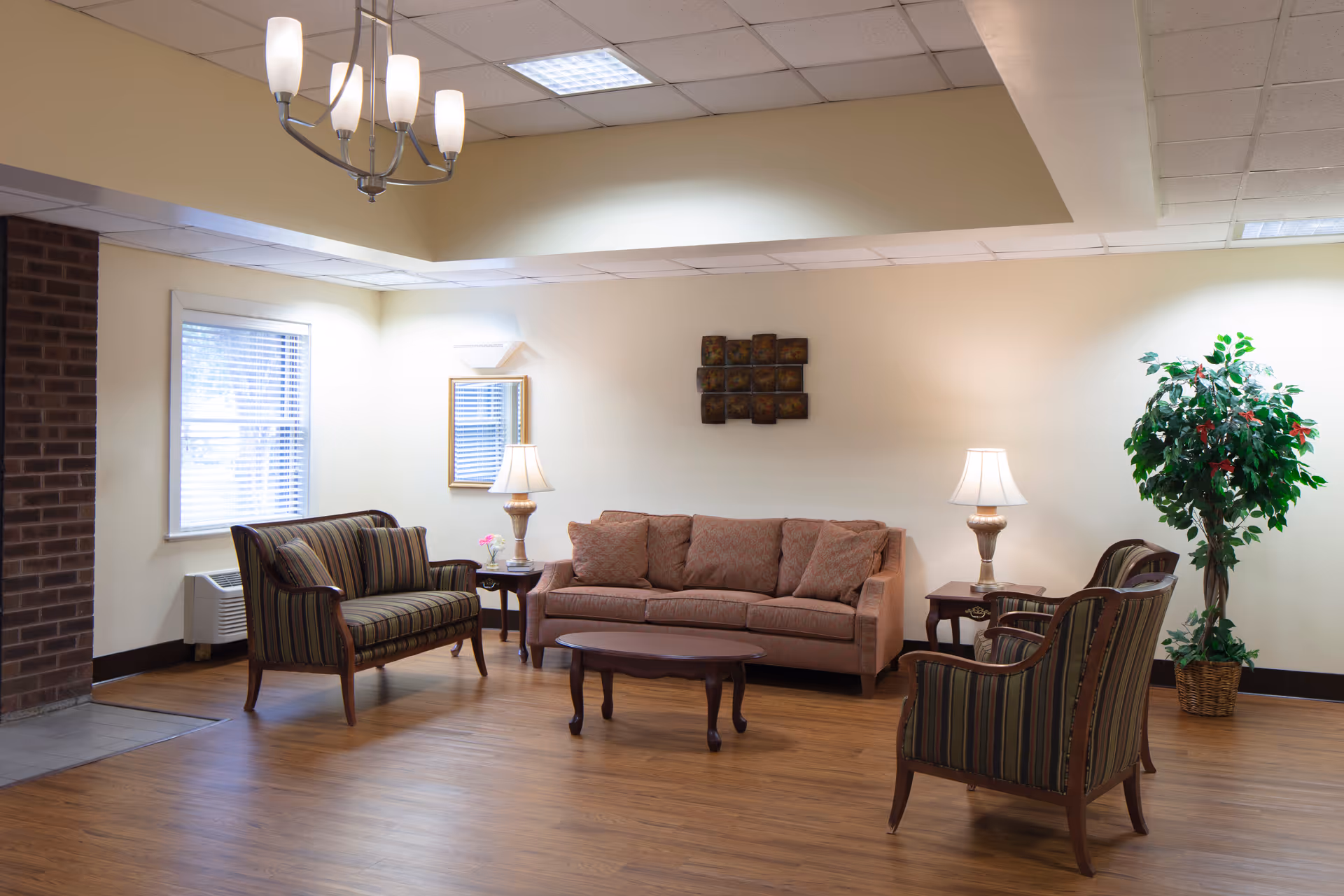 A cozy senior living lounge with sofa, striped chairs, side tables, lamps, and plants.