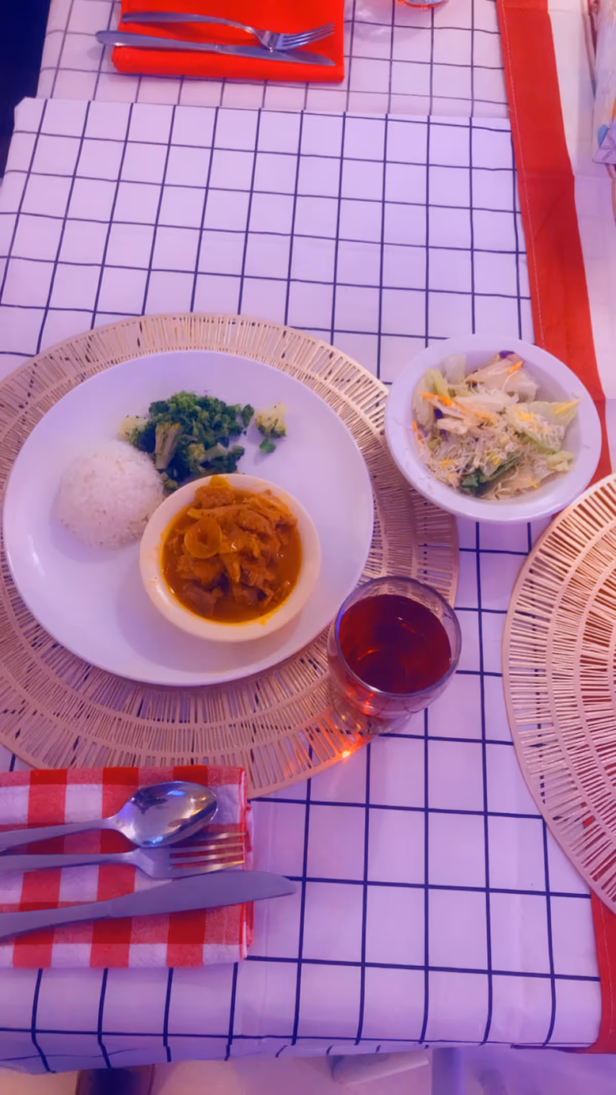 Plate with rice, broccoli, curry, and salad beside a red drink on a checkered tablecloth.