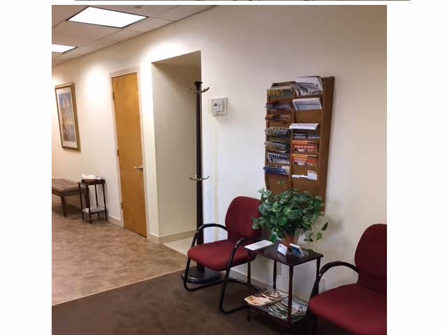 Hallway seating area in a senior living facility with chairs, plants, and a magazine rack.