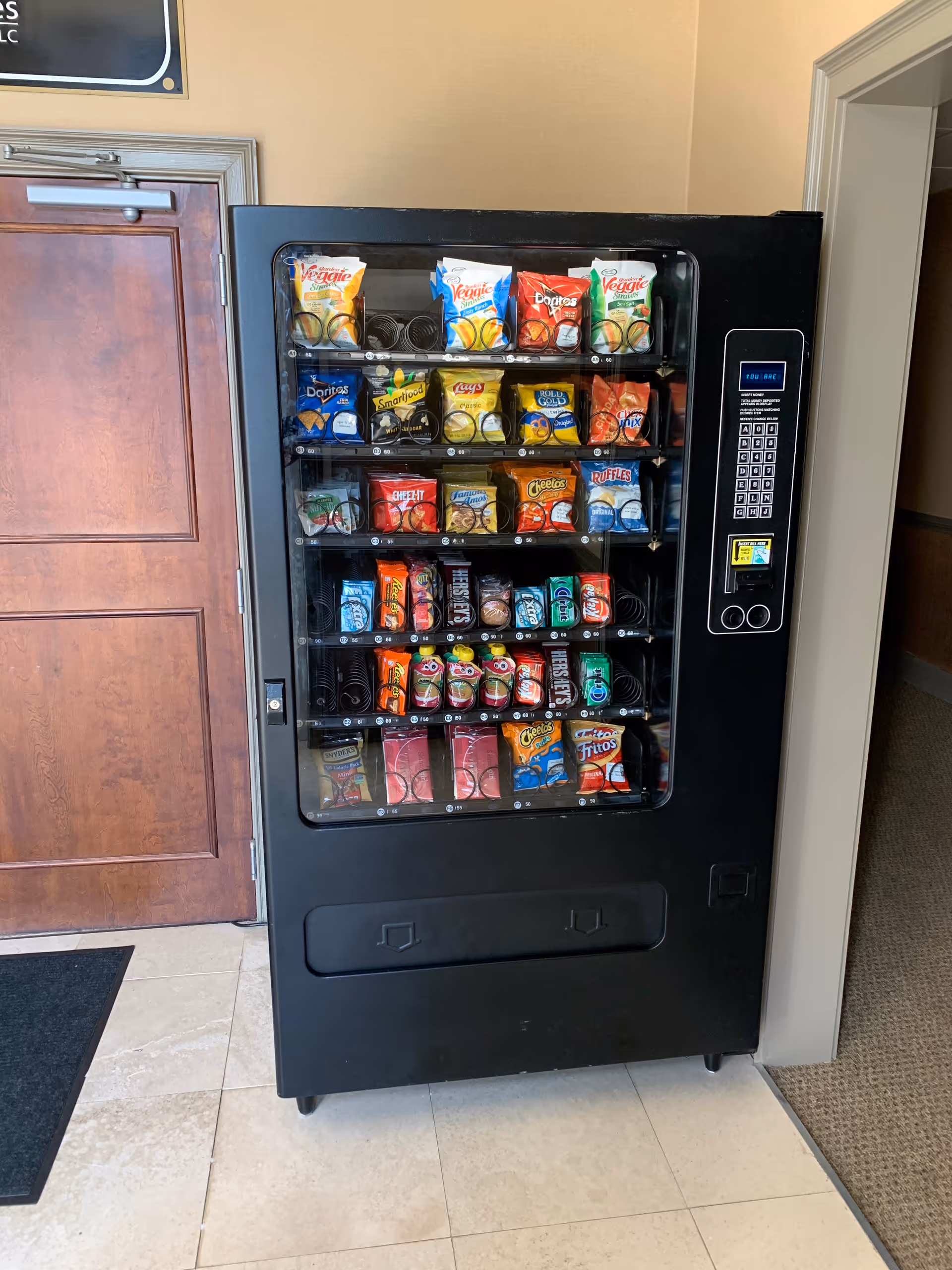 Black snack vending machine stocked with chips inside a beige-walled facility lobby.