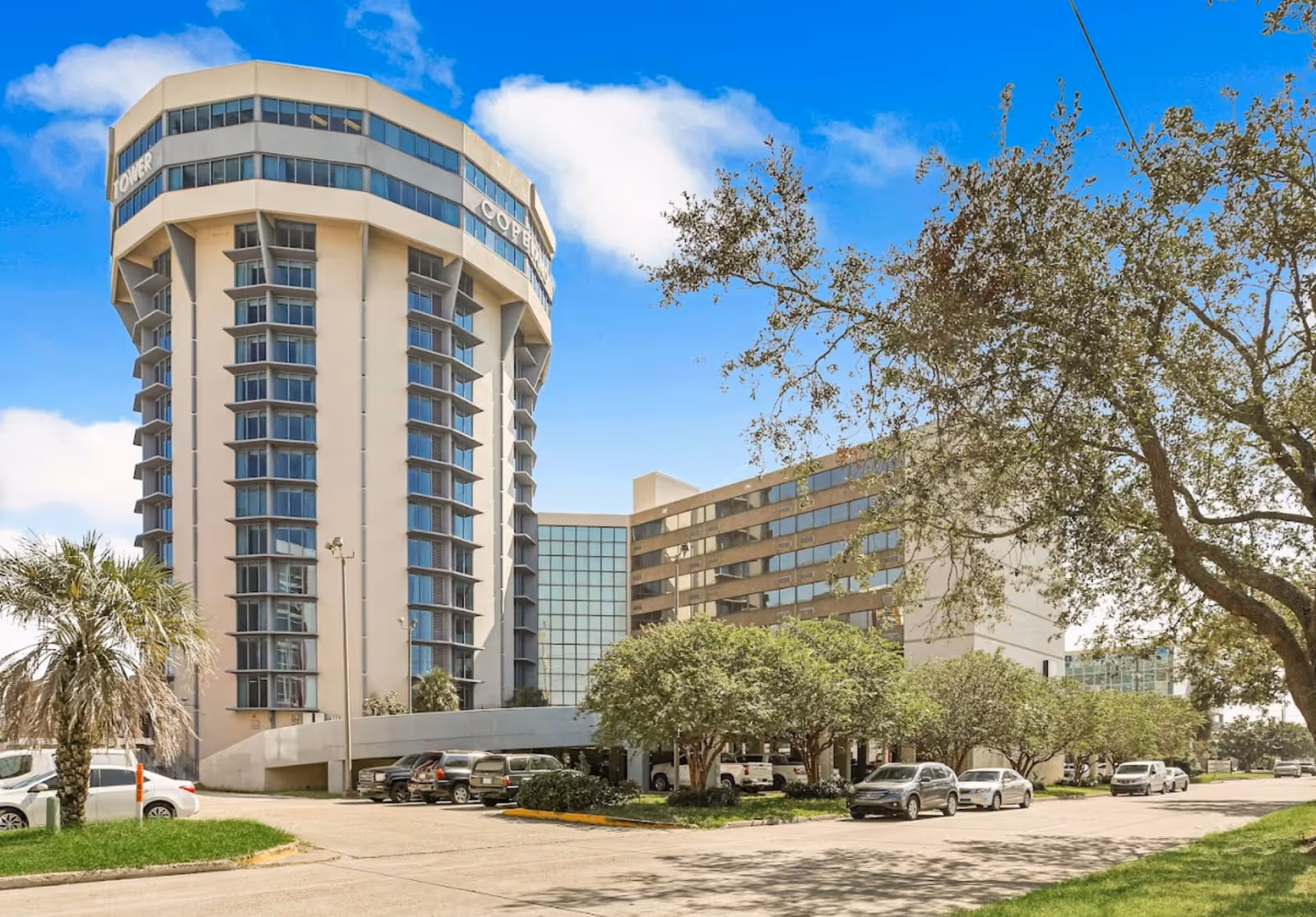 Exterior view of Copeland Tower Living, a tall multi-story building with large windows and a curved structure, surrounded by trees and parked cars under a blue sky with some clouds.