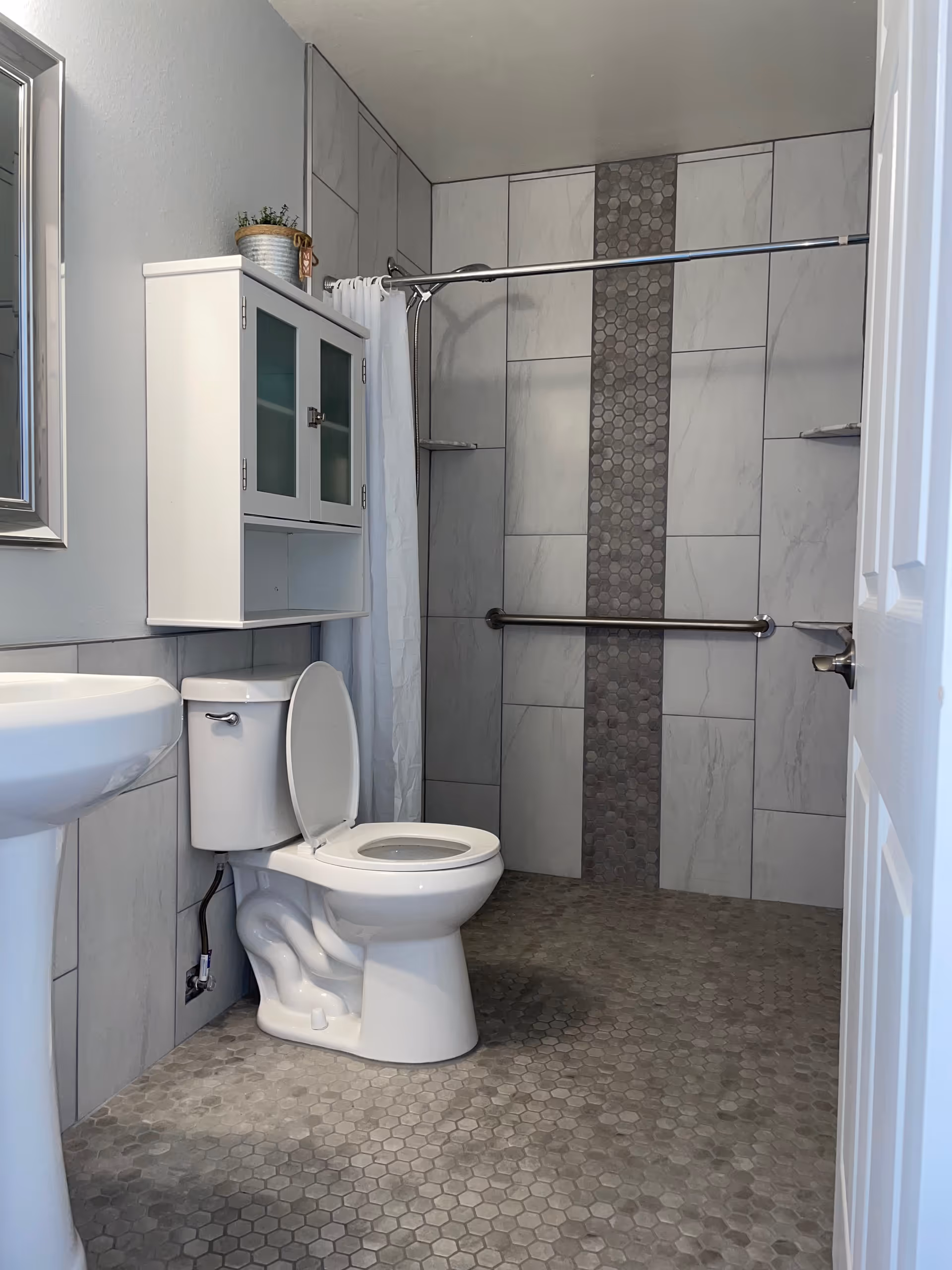 A bathroom with toilet, sink, cabinet, shower, and accessibility grab bars.