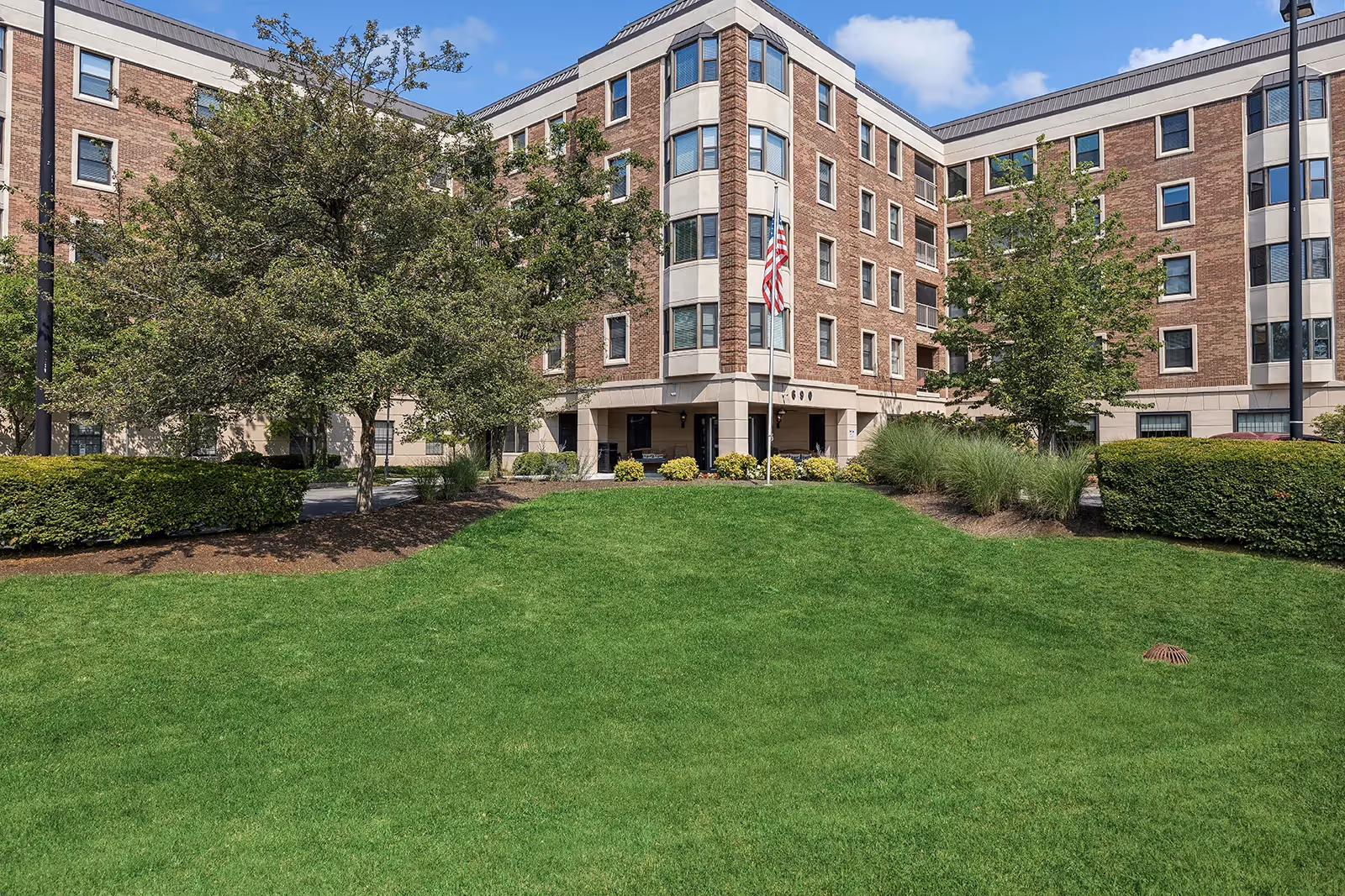 Brick senior living building with manicured lawn, trees, and an American flag near entrance.